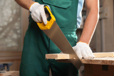 Photo of Professional carpenter cutting wooden board with handsaw in workshop, closeup
