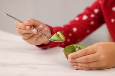 Girl eating tasty fresh kiwi with spoon at white marble table indoors, closeup