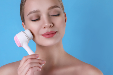 Young woman washing face with cleansing brush on light blue background. Cosmetic product