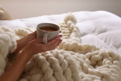 Woman covered with knitted plaid holding cup of tea in bed, closeup