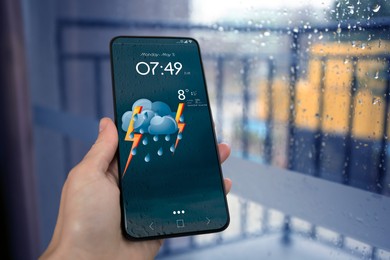Woman checking weather using app on smartphone near wet window, closeup. Data and illustration of cloud with lightning and rain on screen