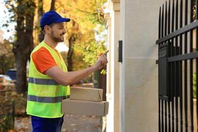 Photo of Courier in uniform with parcels ringing doorbell outdoors