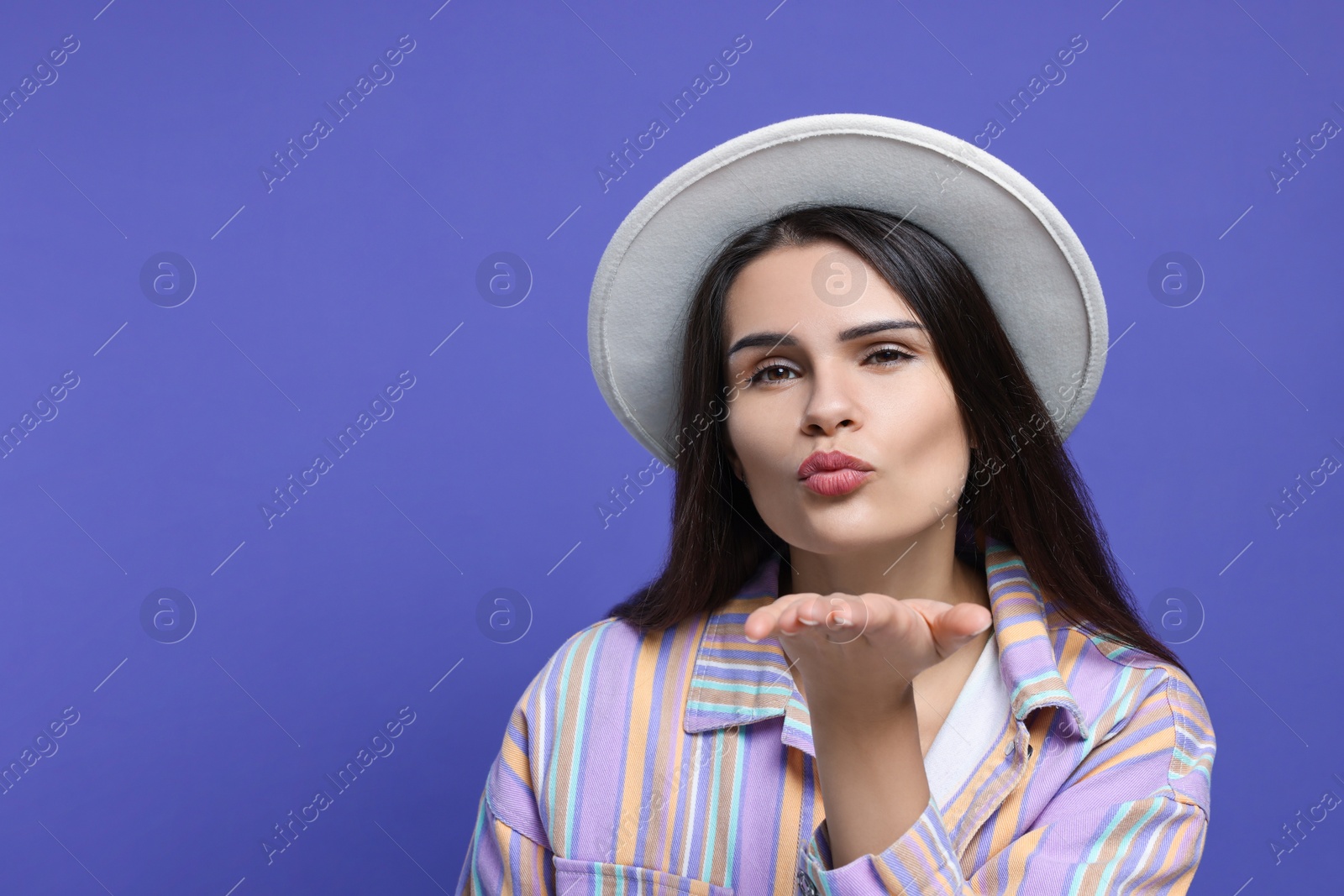 Photo of Beautiful young woman with stylish hat blowing kiss on purple background. Space for text