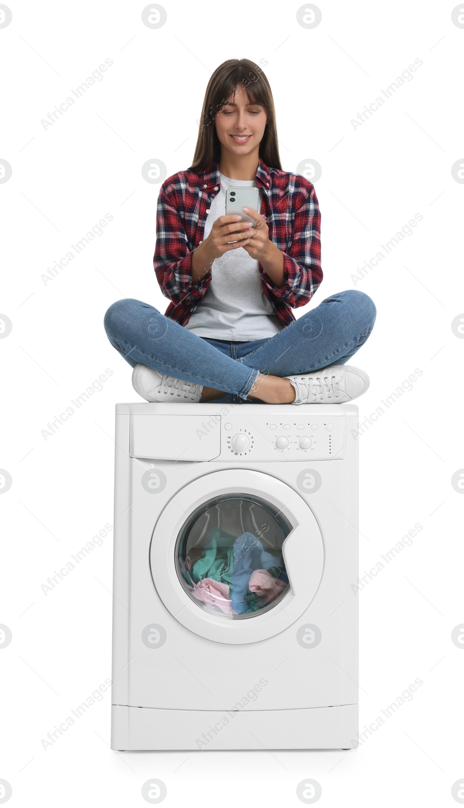 Photo of Beautiful woman using smartphone while sitting on washing machine with laundry against white background