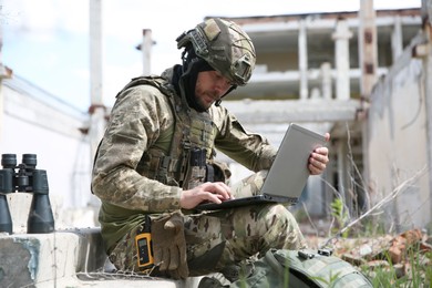 Photo of Military mission. Soldier in uniform using laptop near abandoned building outside