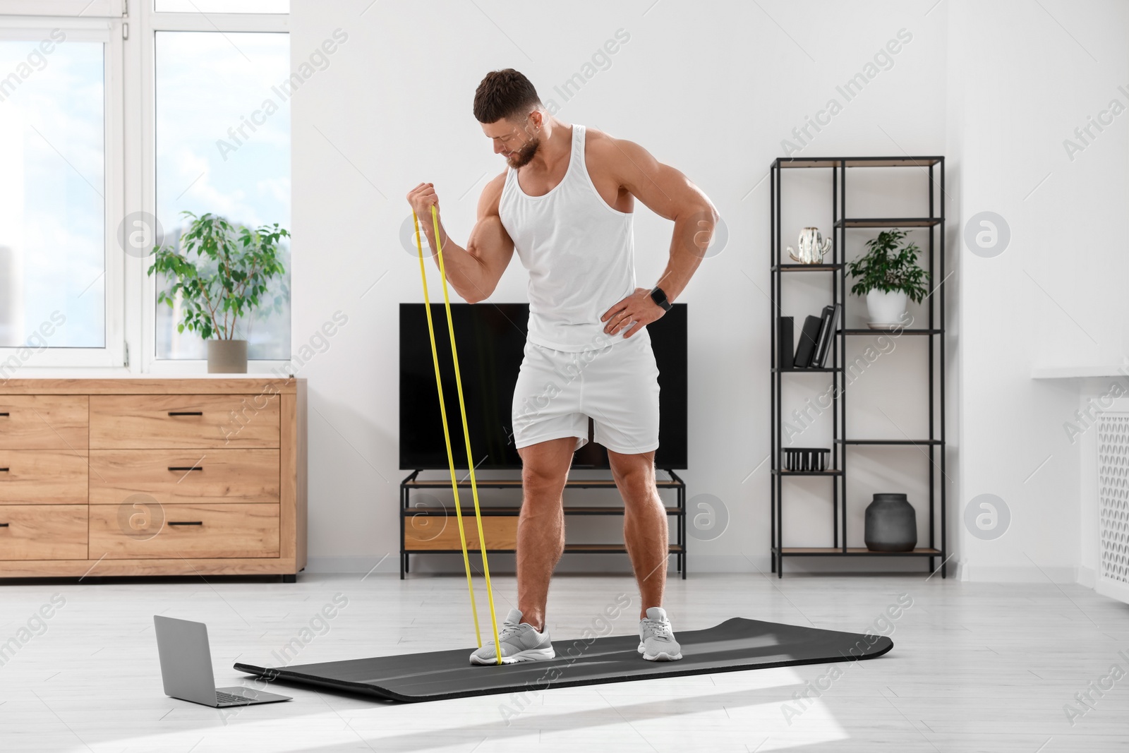 Photo of Muscular man doing exercise with elastic resistance band near laptop on mat at home