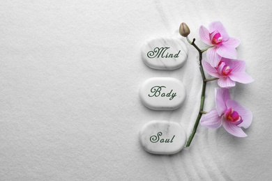 Stones with words Mind, Body, Soul and flowers on sand, flat lay. Zen lifestyle