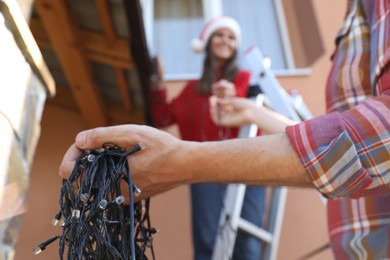 Woman in Santa hat and man decorating house with Christmas lights outdoors, selective focus