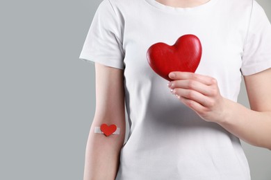 Blood donation concept. Woman with adhesive plaster on arm holding red heart against grey background, closeup. Space for text