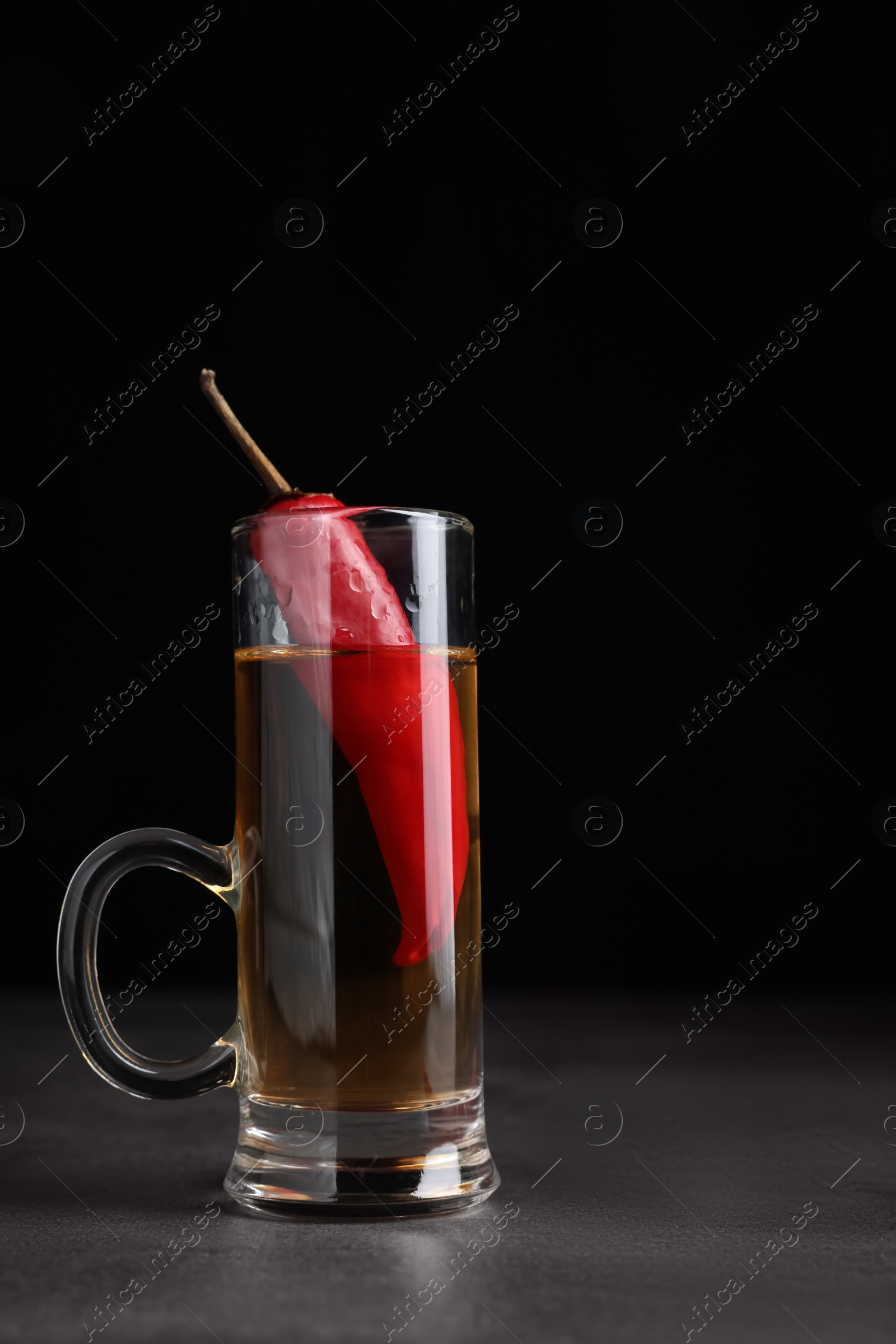 Photo of Red hot chili peppers and vodka in shot glass on grey table against black background