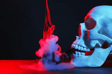 Human skull with pipe and smoke in neon lights on black background, closeup. Space for text