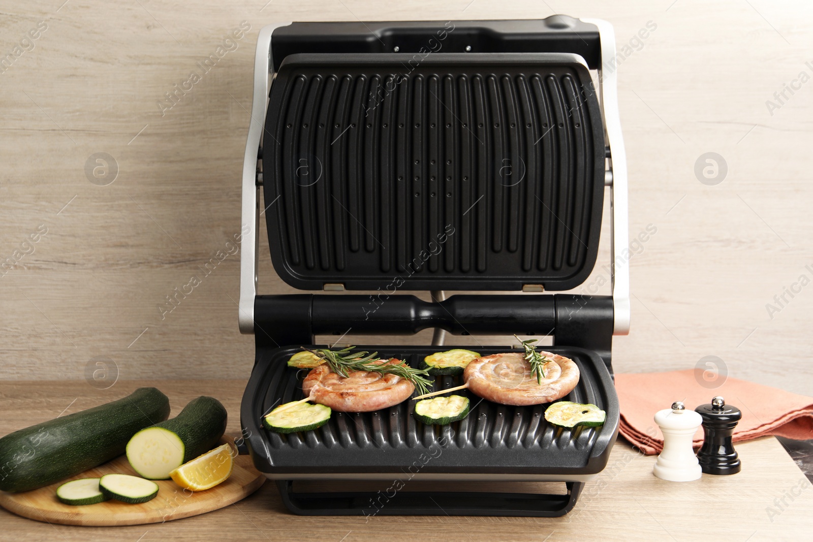 Photo of Electric grill with homemade sausages, zucchini and rosemary on wooden table