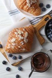 Photo of Delicious croissants with almond flakes, chocolate and blueberries served on white wooden table, flat lay