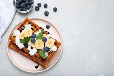 Photo of Plate of delicious Belgian waffles with blueberry, banana, whipped cream and chocolate sauce on light marble table, flat lay. space for text