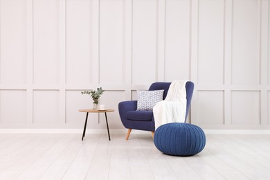 Stylish room interior with comfortable armchair and knitted pouf near white wall