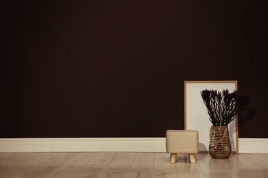 Stylish beige pouf, poster and decorative flowers near brown wall in room. Space for text