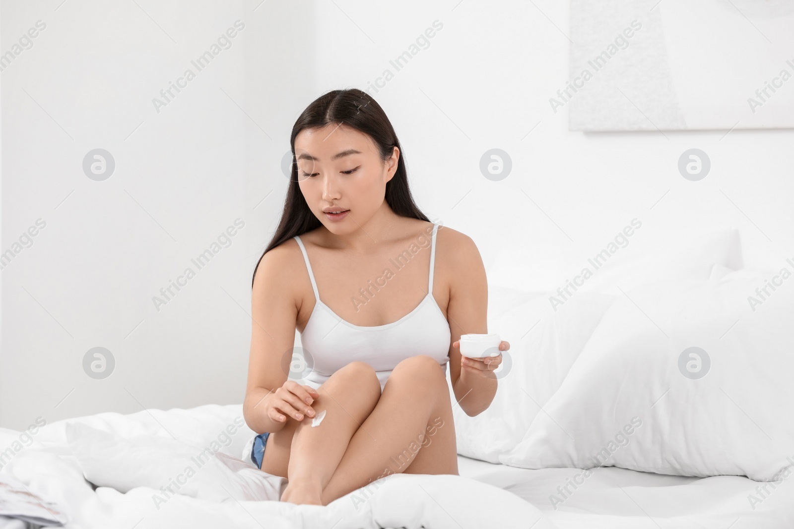 Photo of Beautiful young Asian woman applying body cream on leg in bedroom