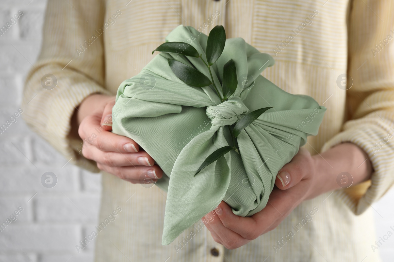 Photo of Furoshiki technique. Woman holding gift packed in green fabric and decorated with ruscus branch against white wall, closeup