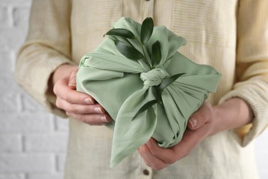 Furoshiki technique. Woman holding gift packed in green fabric and decorated with ruscus branch against white wall, closeup