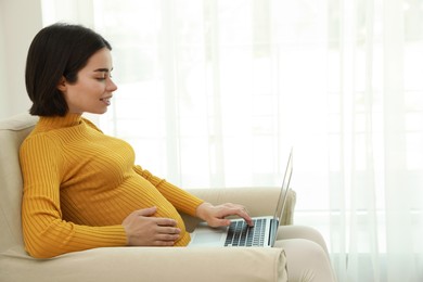 Pregnant young woman with laptop at home, space for text