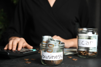 Photo of Woman using calculator near glass jar with money and tag BUSINESS on black table, closeup