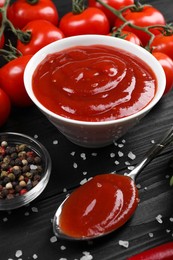 Photo of Delicious ketchup in bowl, spices and tomatoes on black wooden table. Tomato sauce