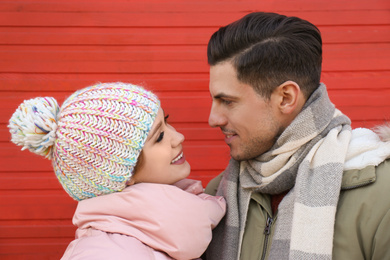 Happy couple in warm clothes near red wooden wall outdoors. Christmas season