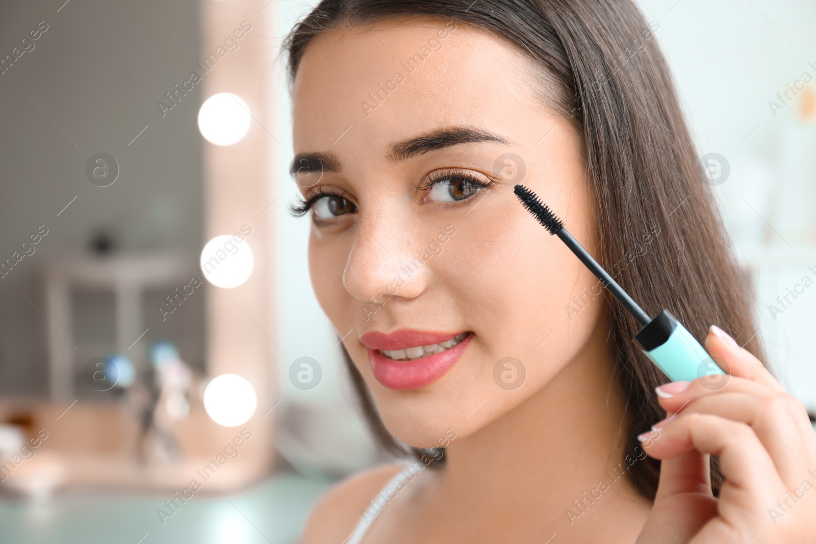 Photo of Attractive young woman applying mascara on her eyelashes against blurred background