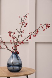 Photo of Hawthorn branches with red berries in vase on wooden table near light wall indoors