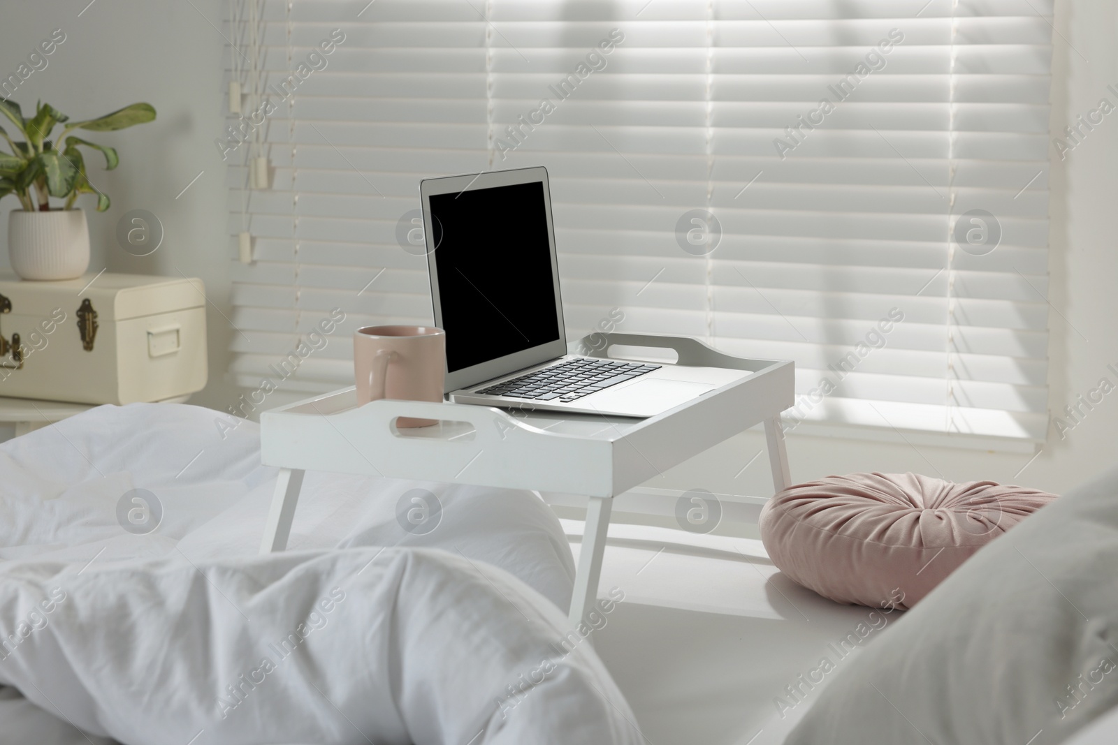 Photo of White tray table with laptop and cup of drink on bed indoors