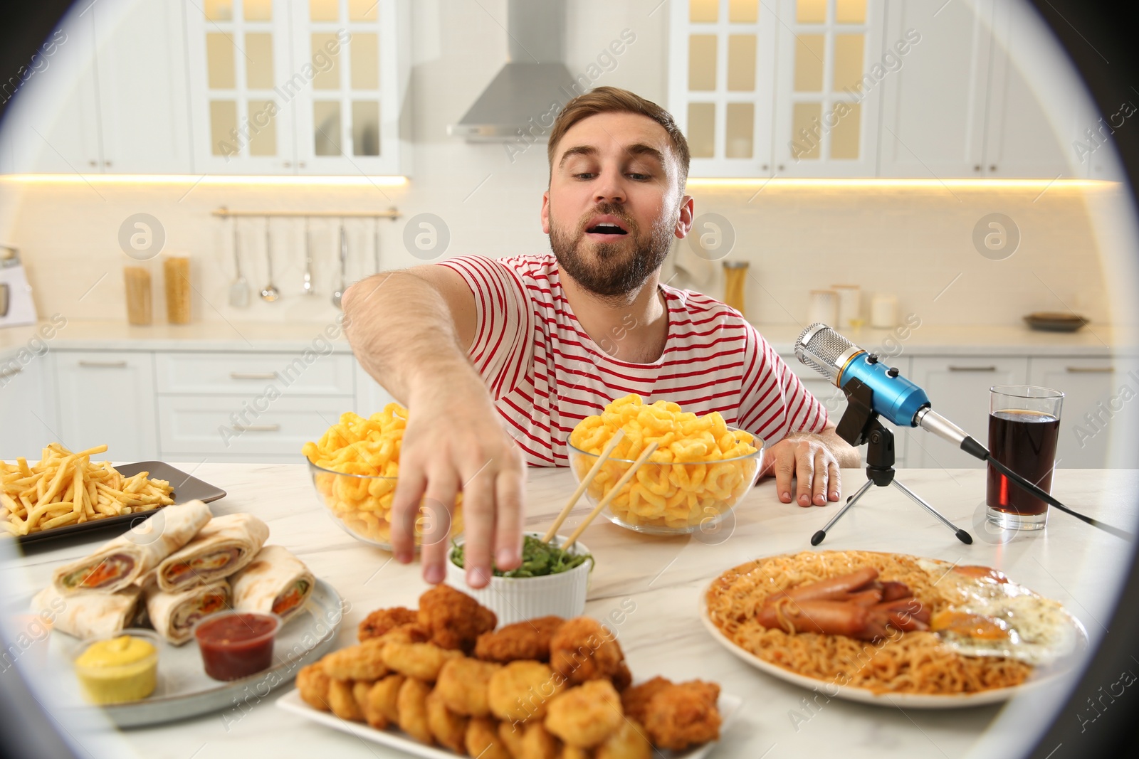 Photo of Food blogger eating near microphone at table in kitchen, view through ring light. Mukbang vlog