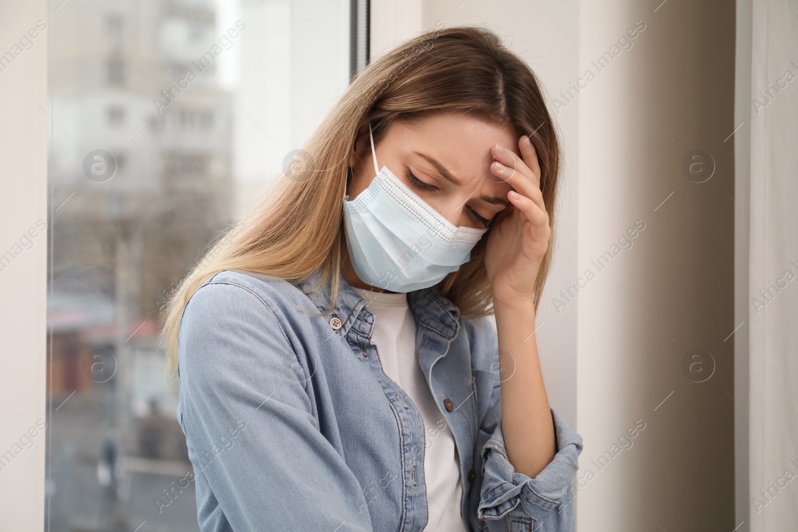 Photo of Sad woman in protective mask near window indoors. Staying at home during coronavirus pandemic