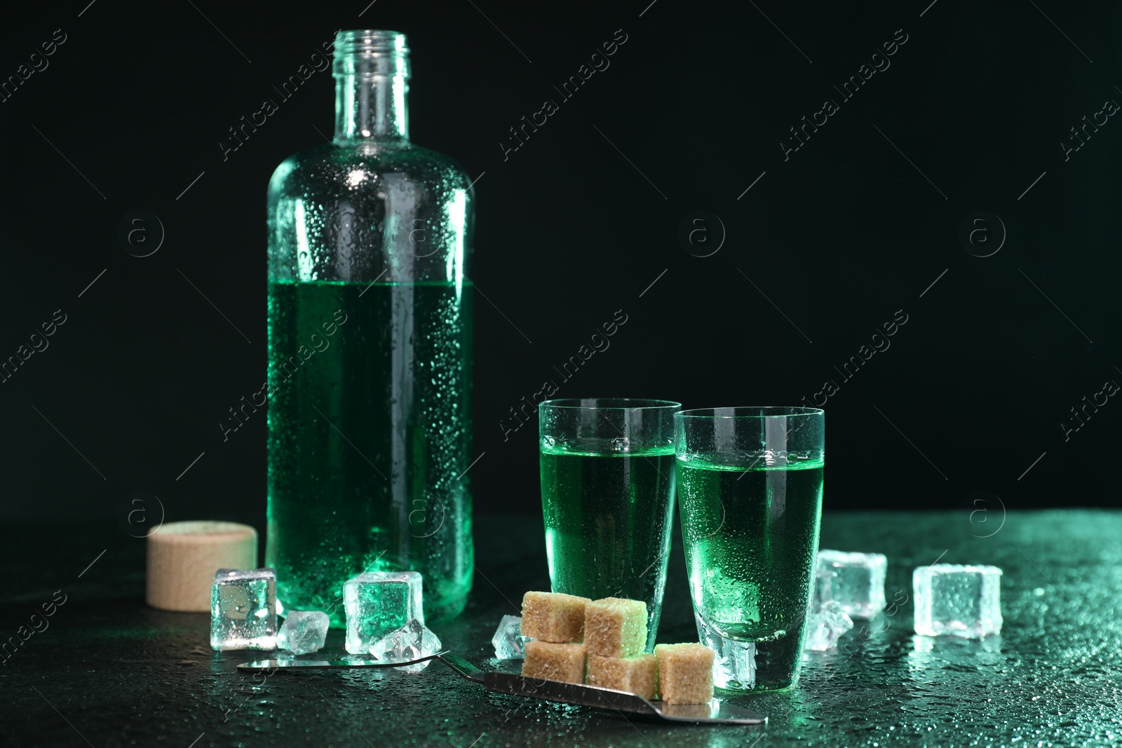 Photo of Absinthe in shot glasses, spoon, brown sugar and ice cubes on gray table against dark background. Alcoholic drink