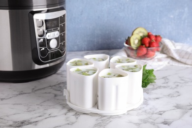 Cups of homemade fruit yogurt near multi cooker on marble table