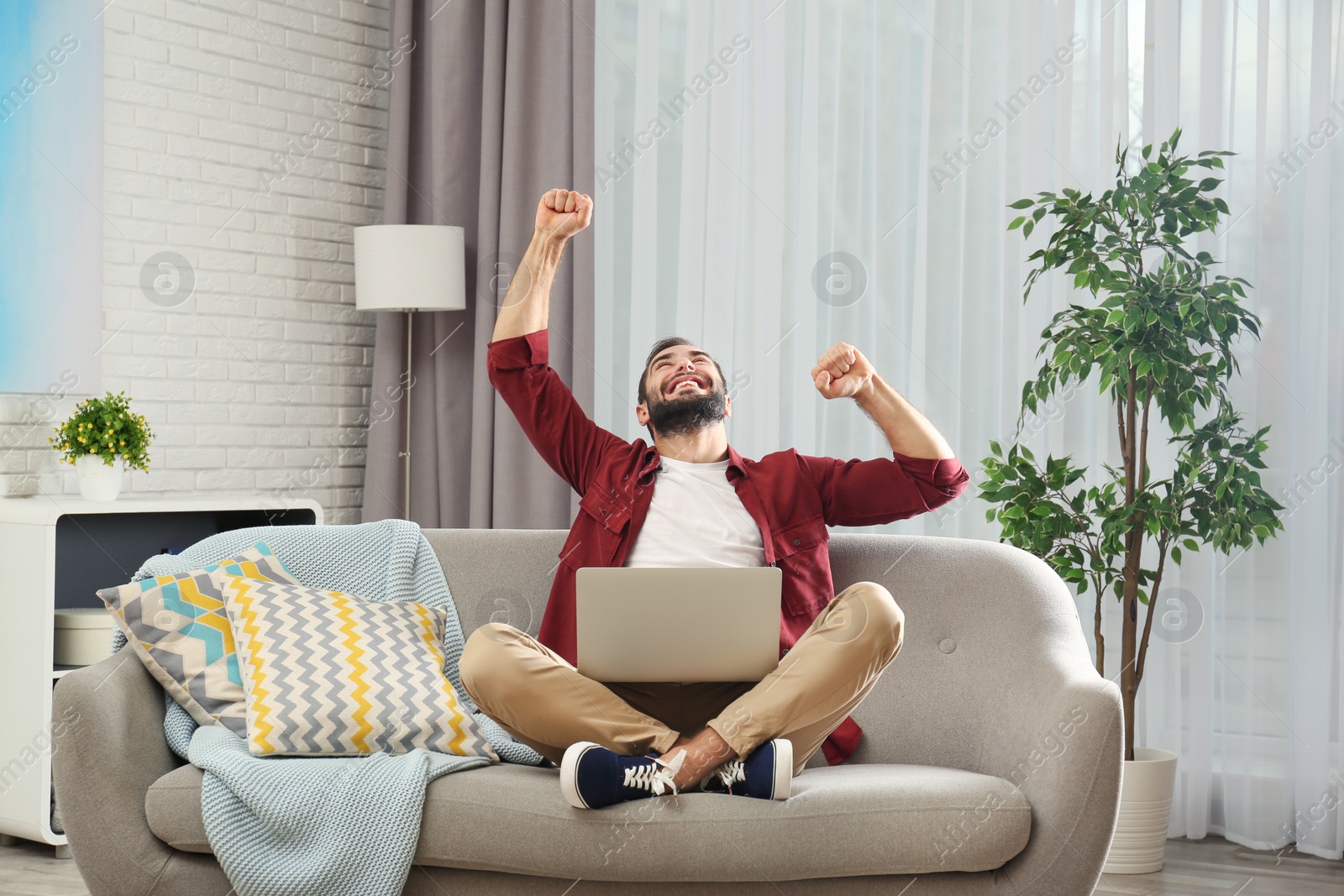 Photo of Emotional young man with laptop celebrating victory on sofa at home