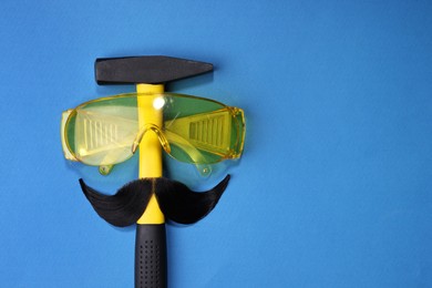 Photo of Man's face made of artificial mustache, safety glasses and hammer on blue background, top view. Space for text