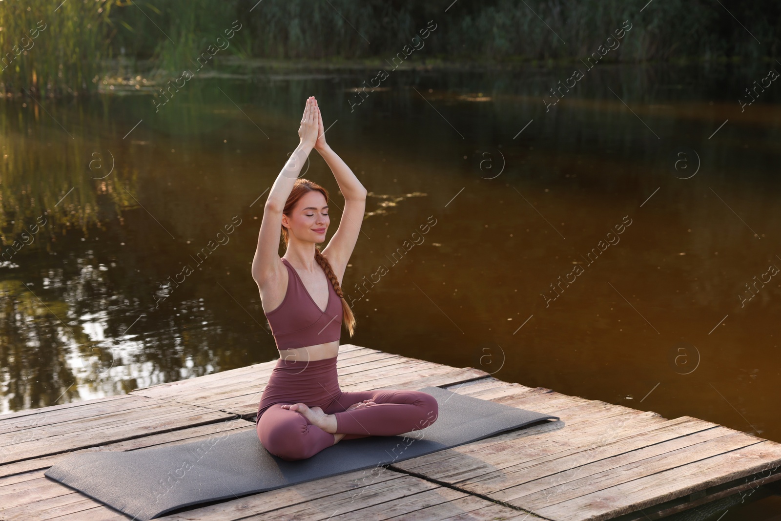 Photo of Beautiful woman practicing Padmasana on yoga mat on wooden pier near pond, space for text. Lotus pose