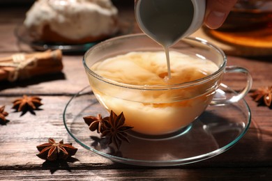 Woman pouring milk into glass cup of tea with anise stars at wooden table, closeup