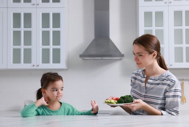 Mother feeding her daughter in kitchen. Little girl refusing to eat vegetables