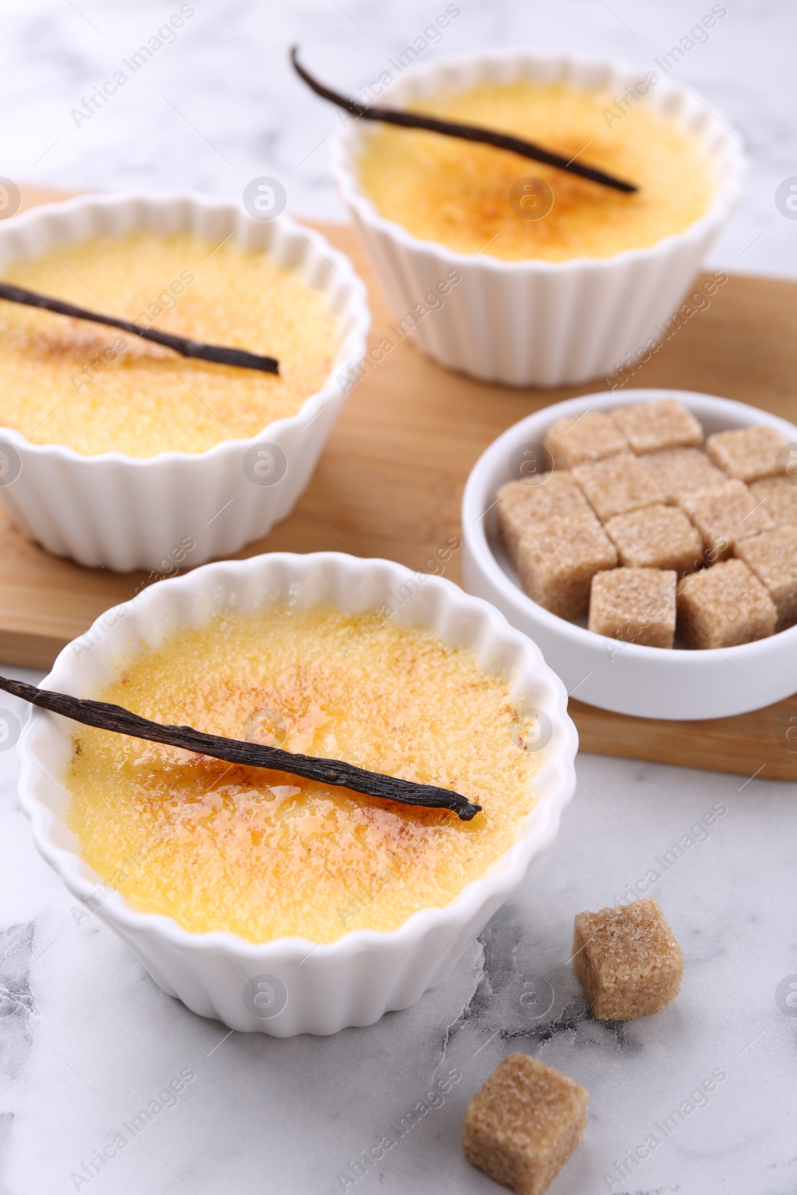 Photo of Delicious creme brulee in bowls, vanilla pods and sugar cubes on white marble table