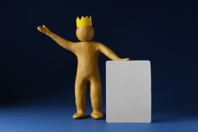 Plasticine figure with crown on head holding blank sign against dark blue background