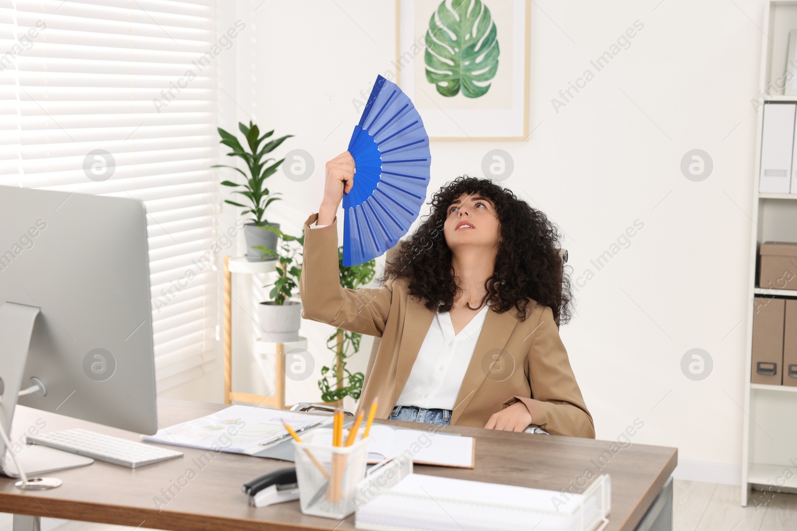 Photo of Young businesswoman waving blue hand fan to cool herself at table in office