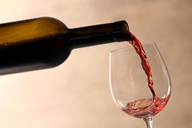 Pouring red wine into glass from bottle against blurred beige background, closeup