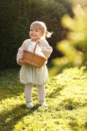 Happy little girl holding wicker basket with cute rabbit outdoors on sunny day
