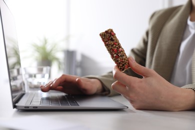 Photo of Woman holding tasty granola bar working with laptop at light table in office, closeup