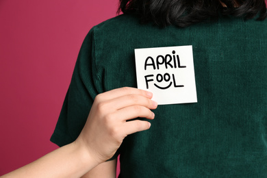 Photo of Woman sticking APRIL FOOL note to friend's back on pink background, closeup