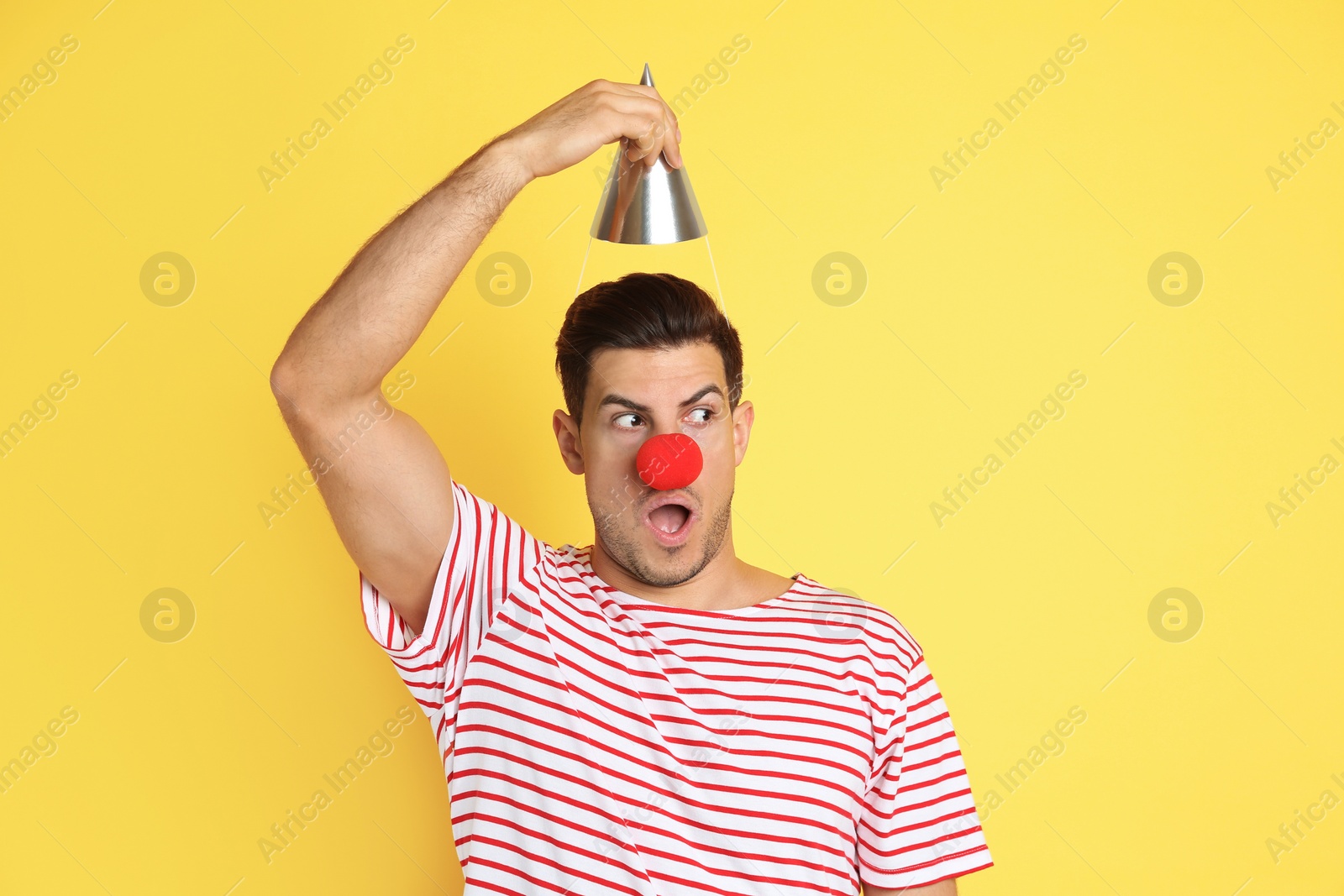 Photo of Funny man with clown nose and party hat on yellow background. April fool's day