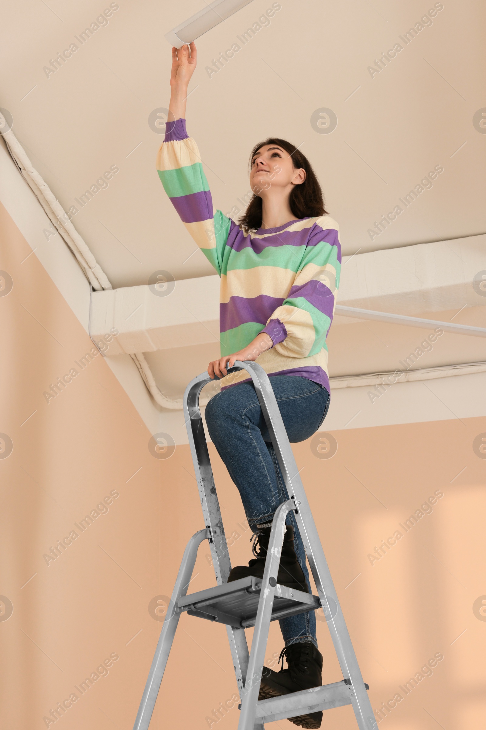 Photo of Young woman installing ceiling lamp on stepladder indoors. Room renovation