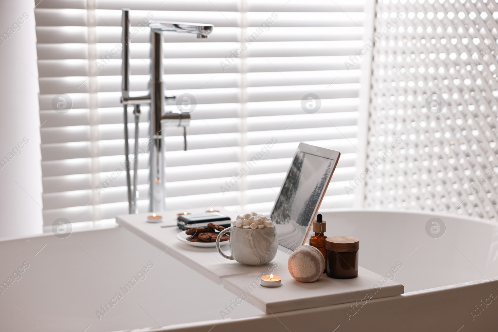 Photo of White wooden tray with tablet, spa products, hot drink and burning candle on bathtub in bathroom
