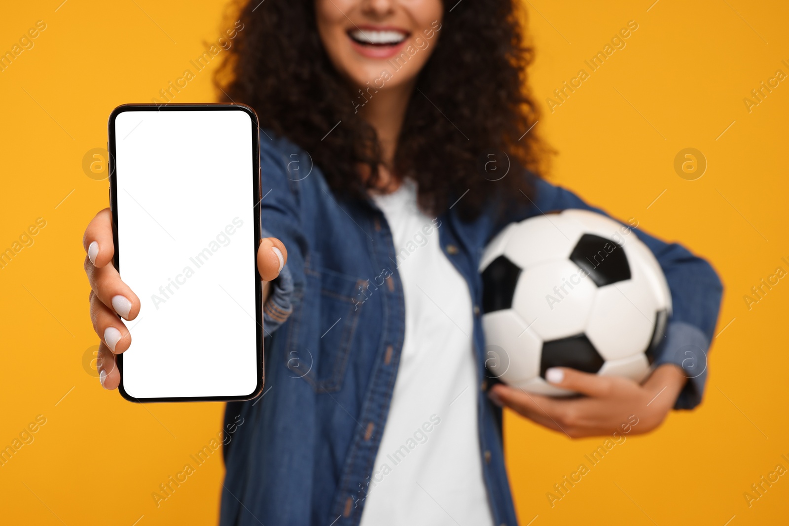 Photo of Happy fan holding soccer ball and showing smartphone on yellow background, closeup